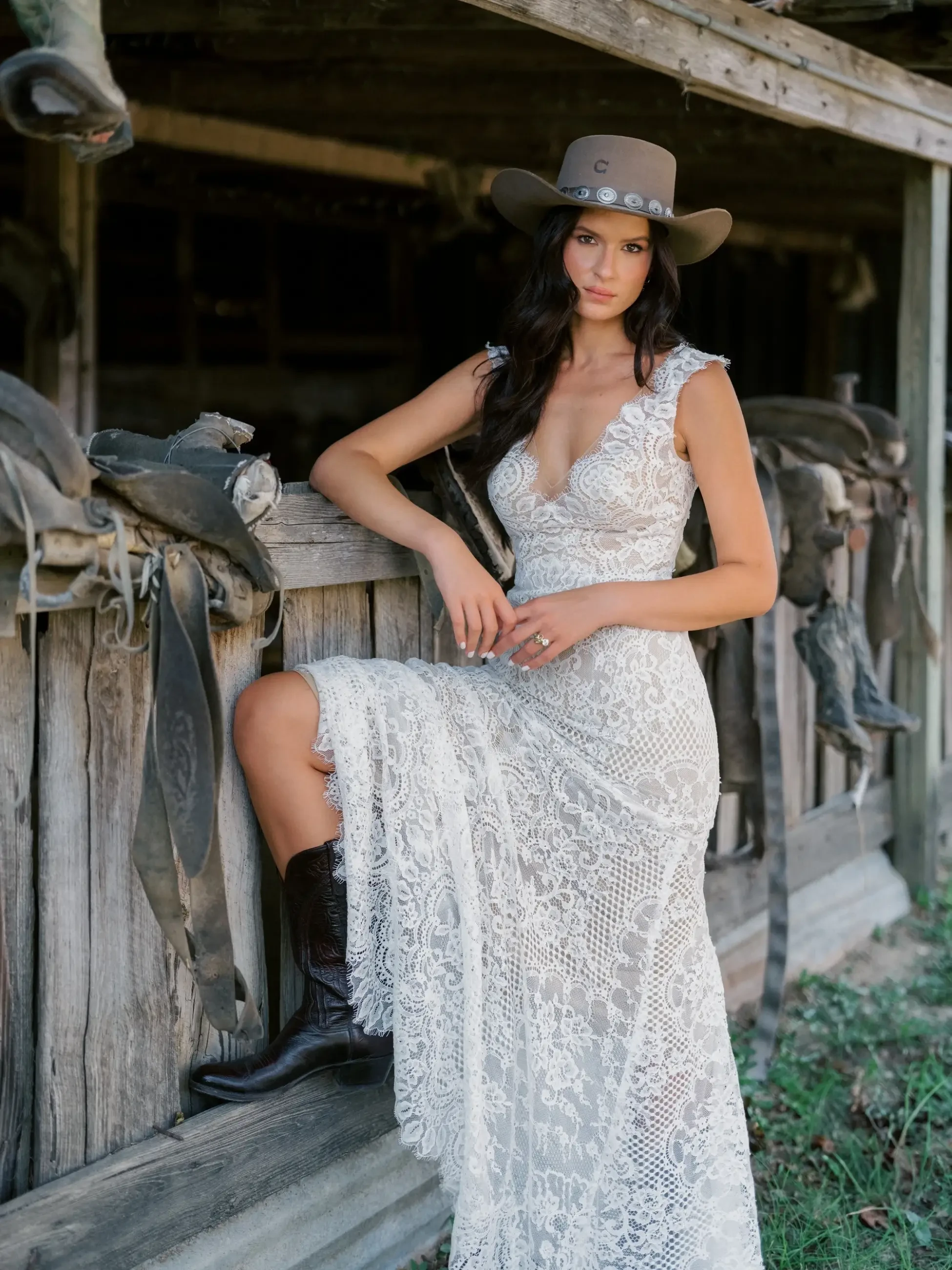 A woman in a white lace dress and cowboy hat poses against a wooden fence with saddles hanging nearby. She is wearing black cowboy boots and has long dark hair.