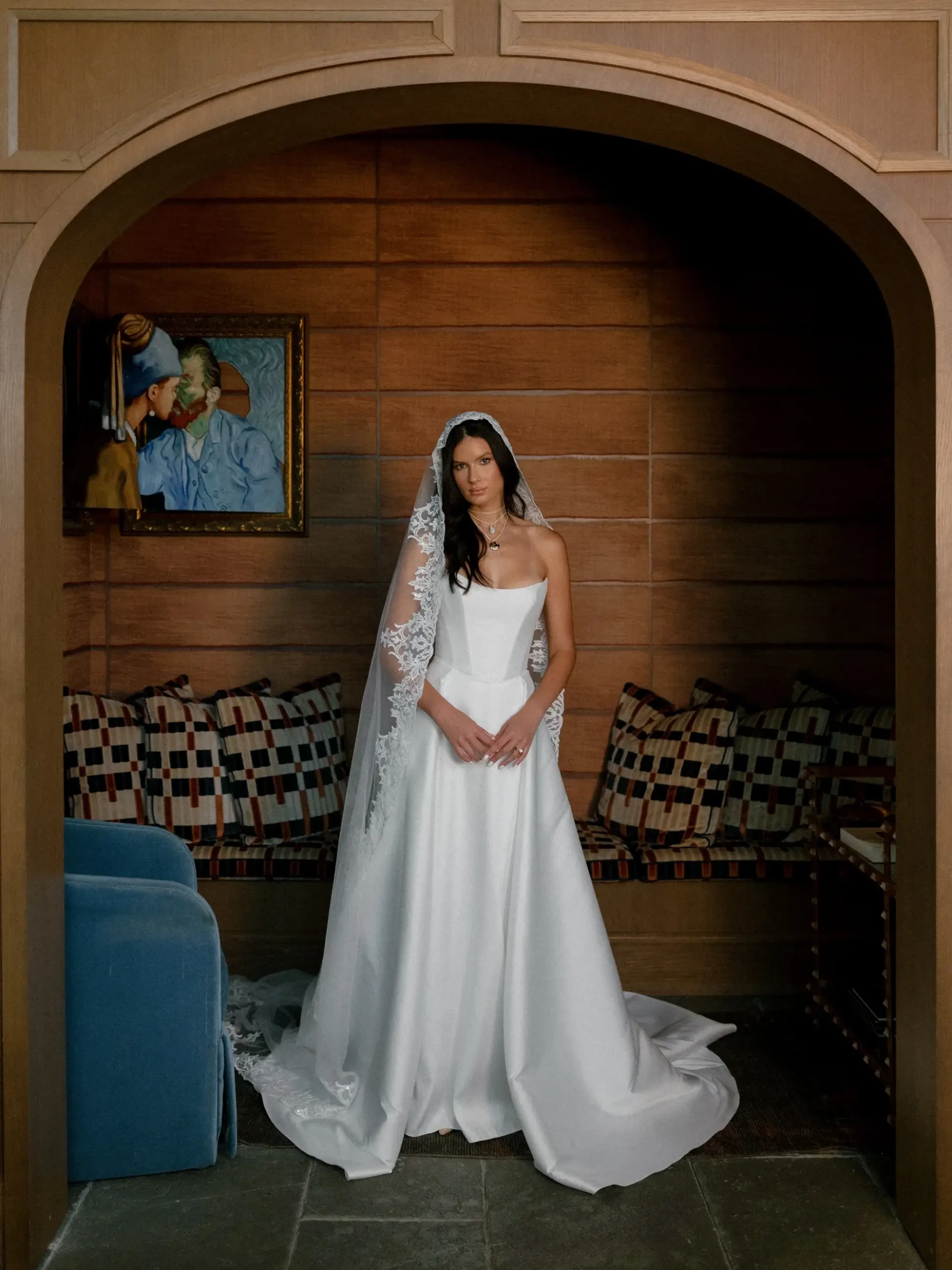Bride in an elegant white dress and lace veil stands under an archway, surrounded by a cozy seating area. A painting hangs on a wooden wall.
