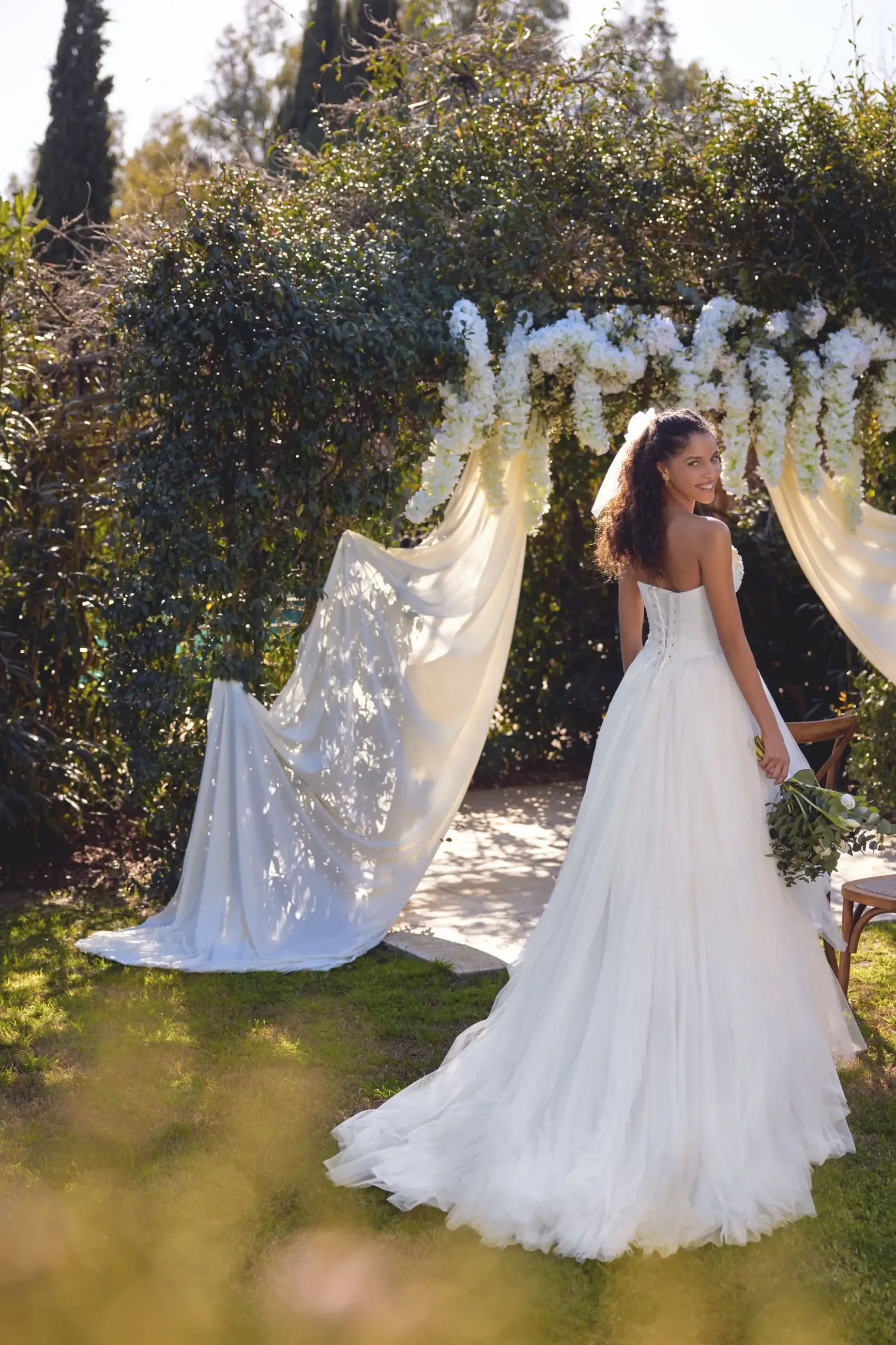 Bride in a flowing white dress stands under a floral arch with sheer drapes in a lush garden, smiling warmly, holding a bouquet.