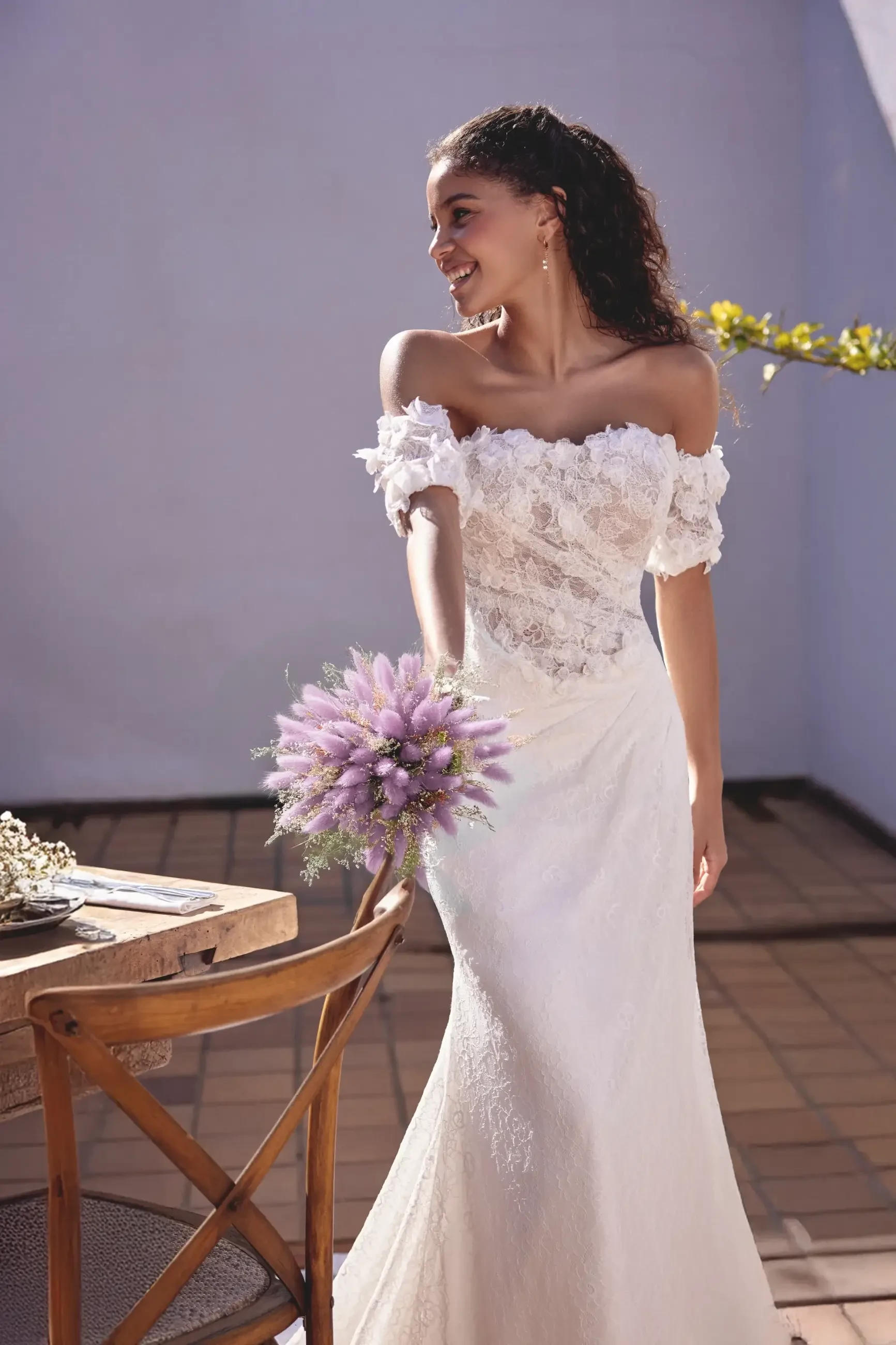 A woman in an off-the-shoulder wedding dress, holding a bouquet of lavender, smiles while standing near a rustic table in a bright, sunny setting.
