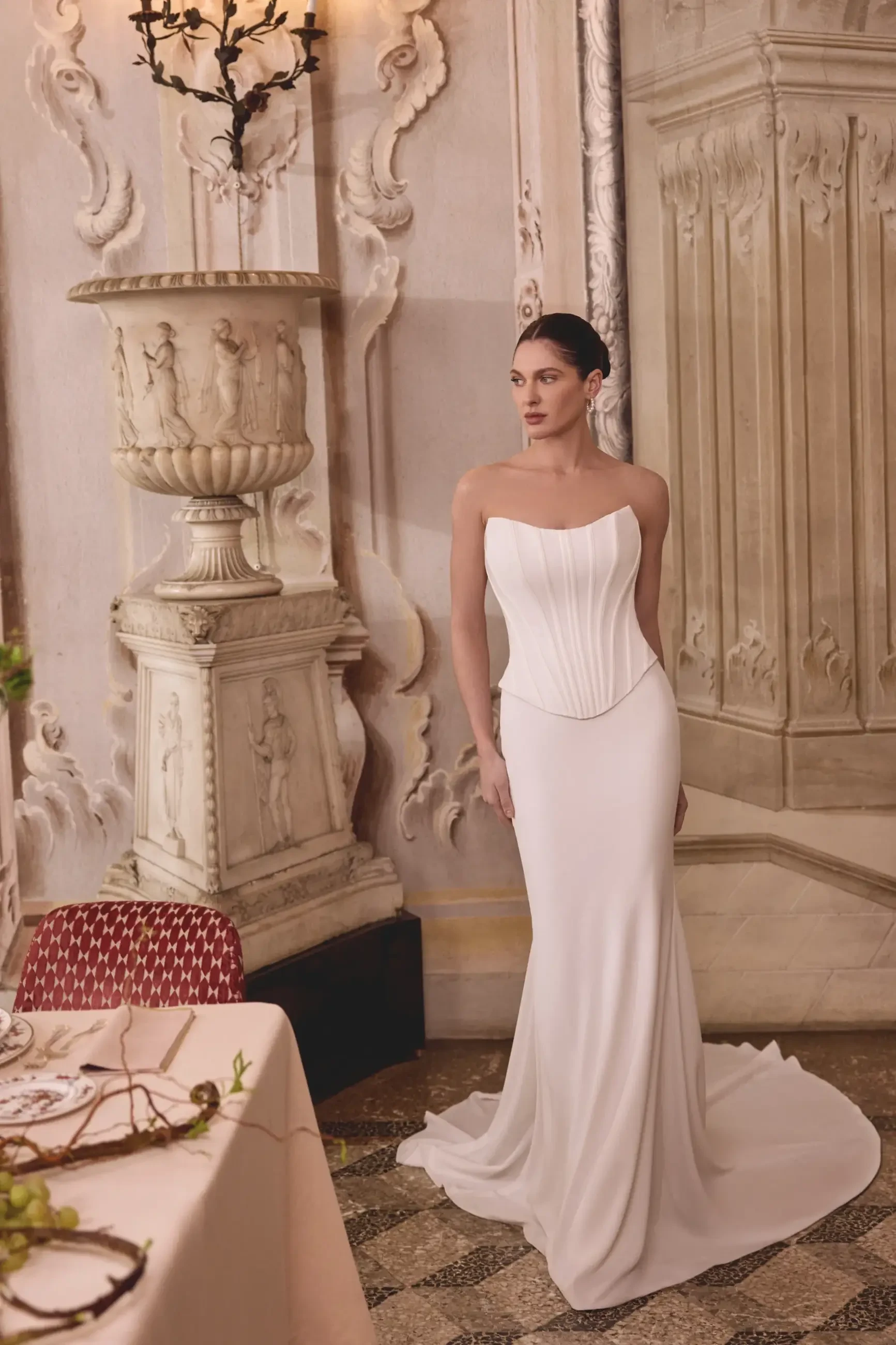 A model wearing a form-fitting white gown with a strapless design, standing elegantly in a beautifully decorated indoor setting.
