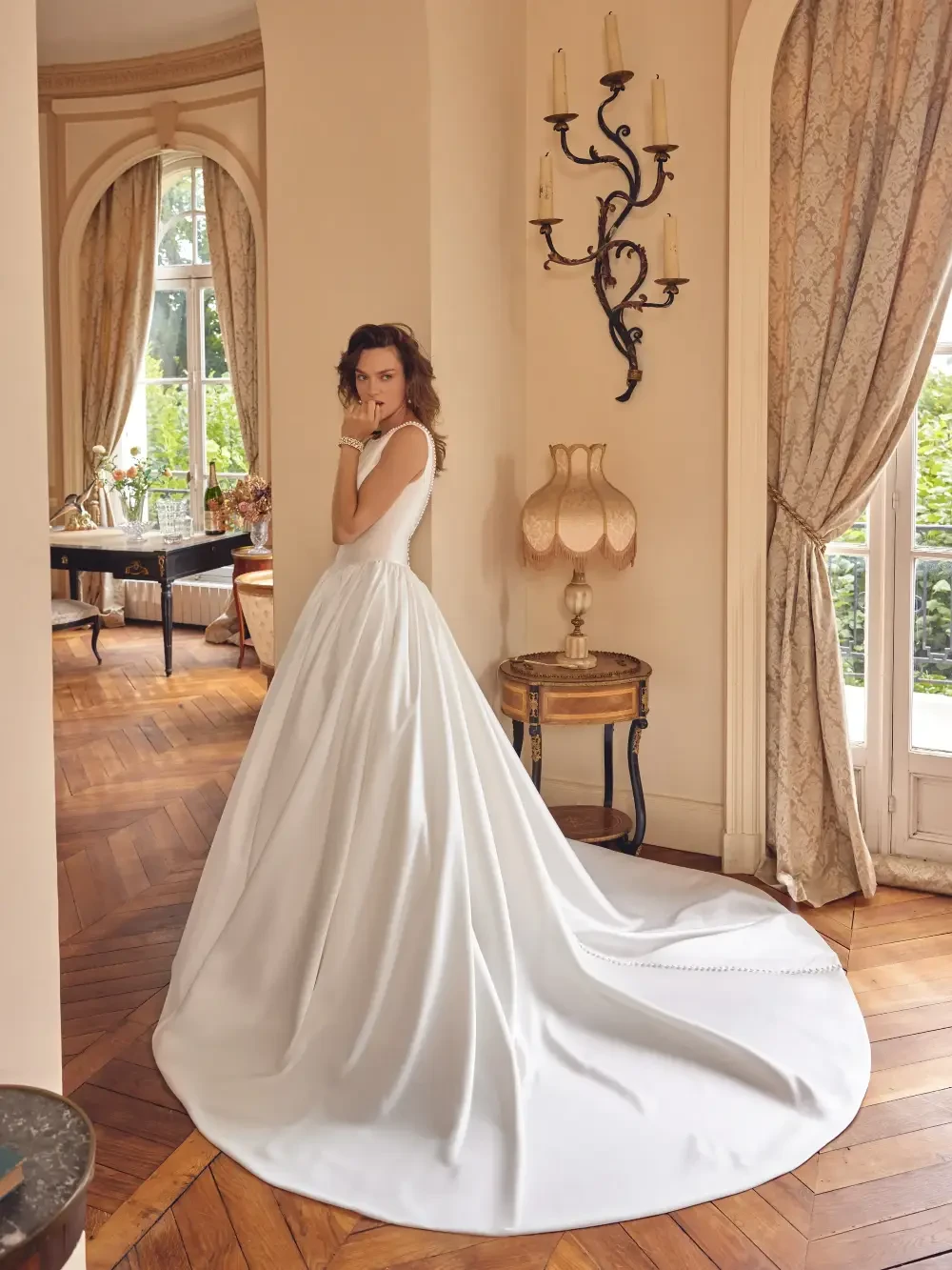A woman in a white wedding dress stands elegantly in a beautifully decorated room with large windows and wooden flooring.