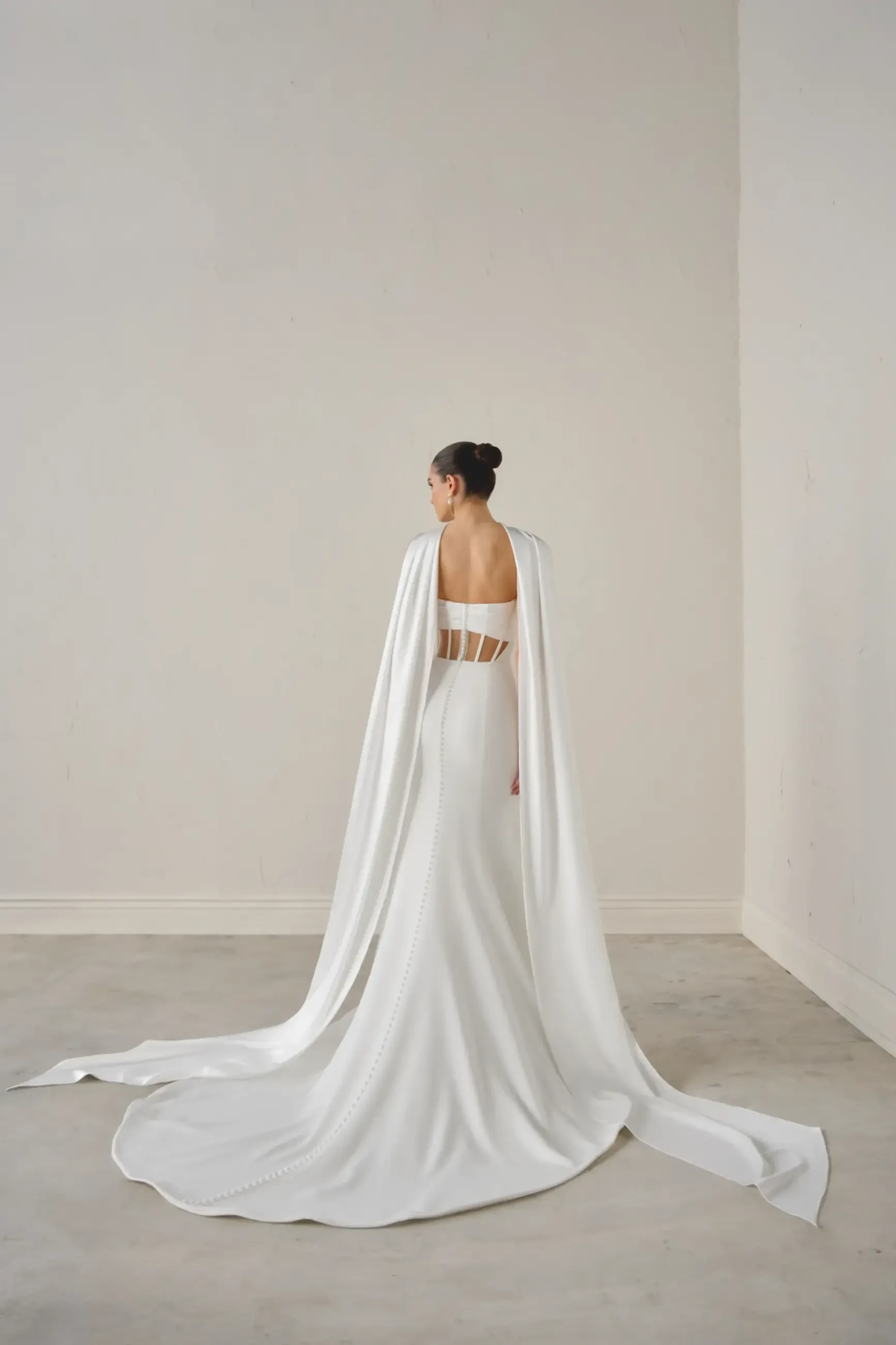 A woman in an elegant white gown with a long, flowing cape stands against a light background. The design is minimalist and sophisticated.