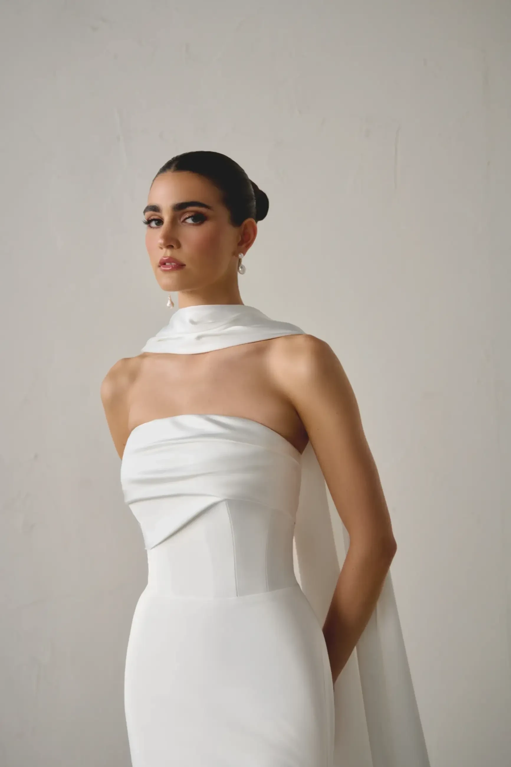 A woman in a sleek, strapless white gown with a flowing scarf around her neck poses against a plain backdrop, exuding elegance and confidence.