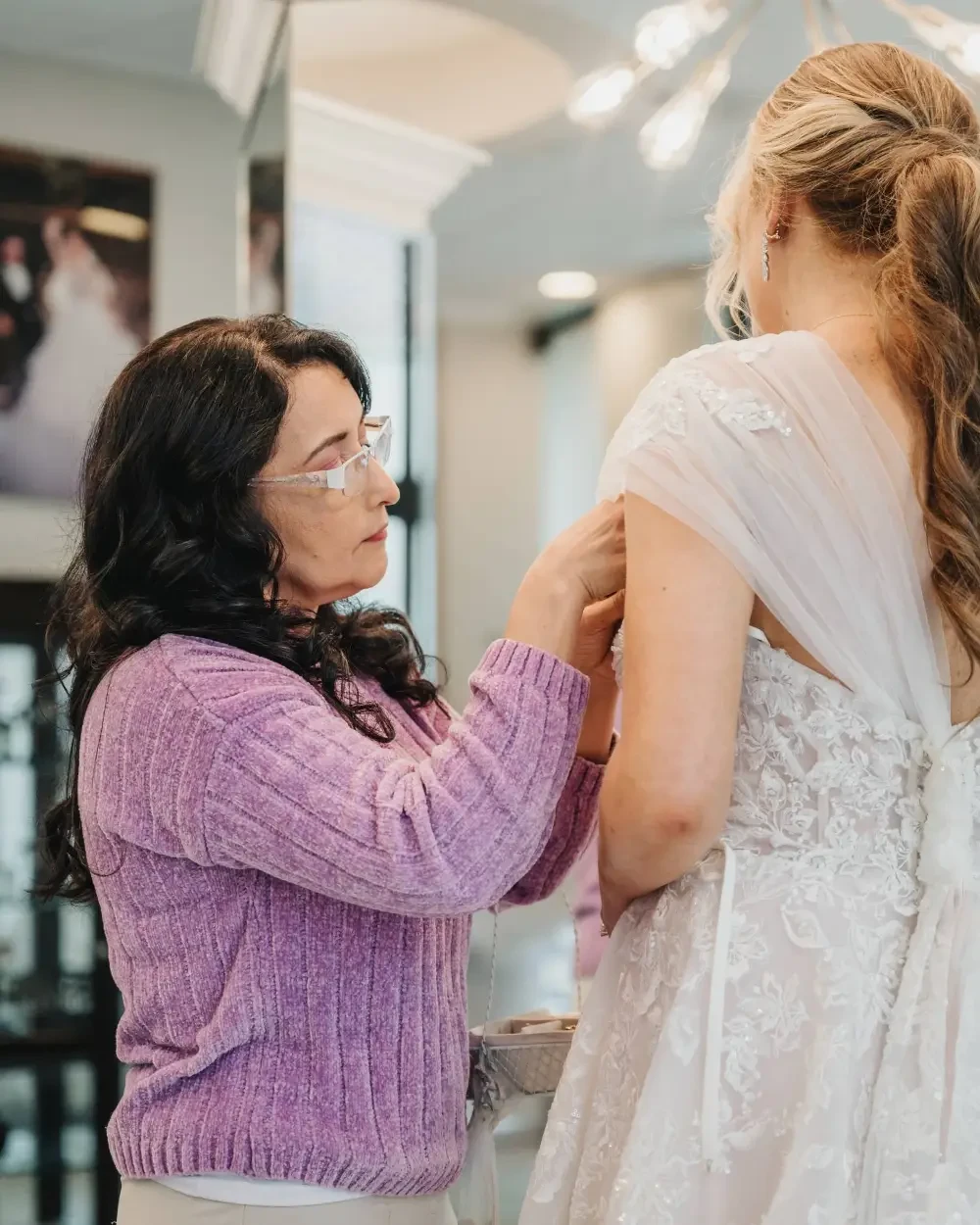 A woman in a wedding dress is being assisted by another woman wearing glasses, in a bridal shop setting.