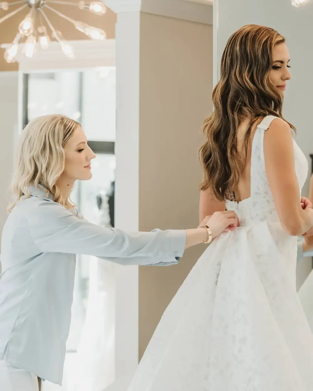 A woman in a wedding dress is having her dress adjusted by a stylist in a bright, modern fitting room.