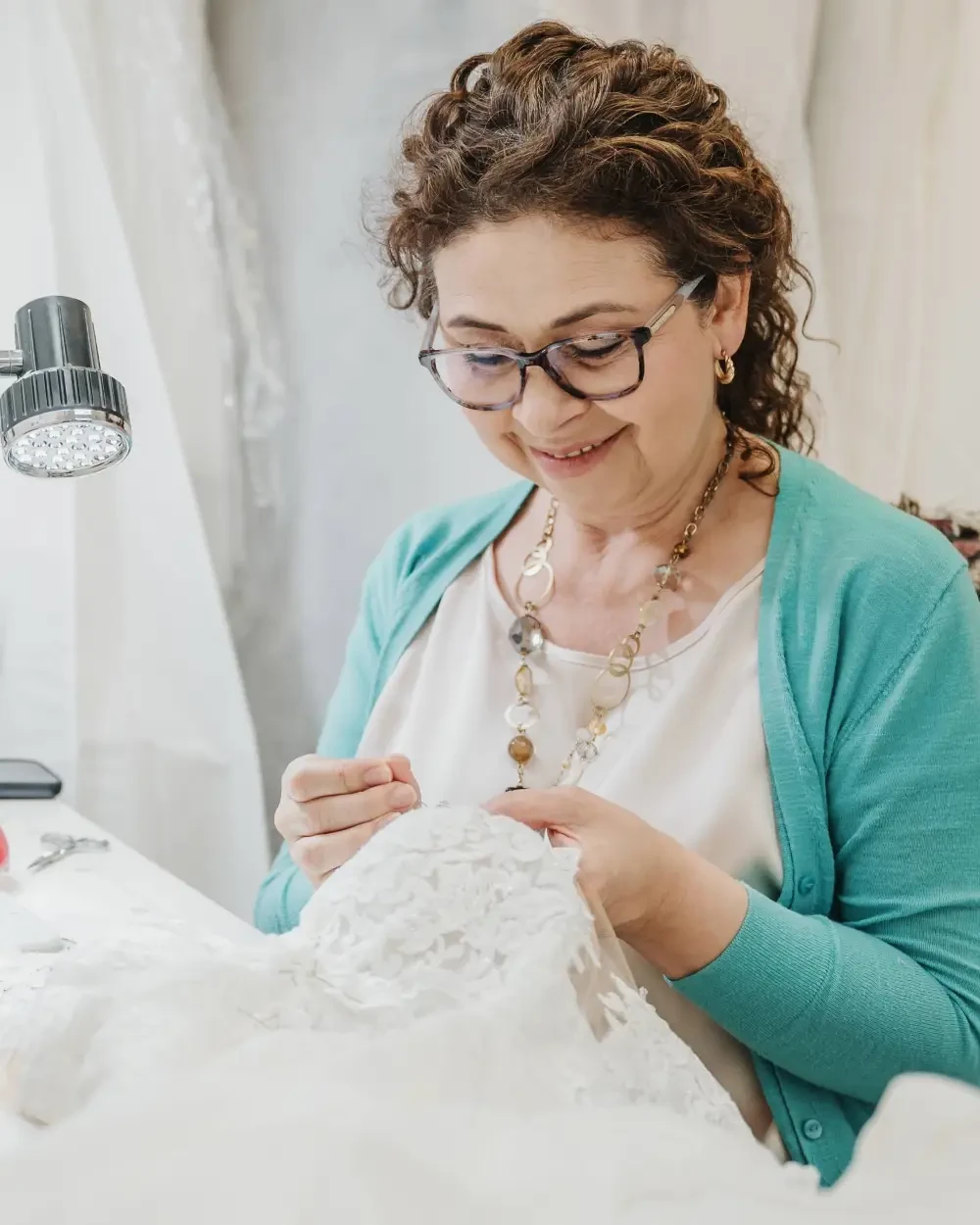 A woman with curly hair and glasses is sewing a piece of lace fabric while seated at a table. She is smiling and appears to be focused on her work.