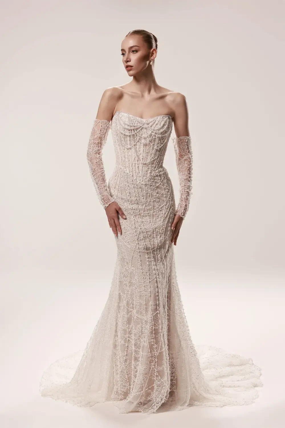 A model wearing an elegant, form-fitting wedding dress with intricate beadwork and sheer long sleeves, posing against a plain background.