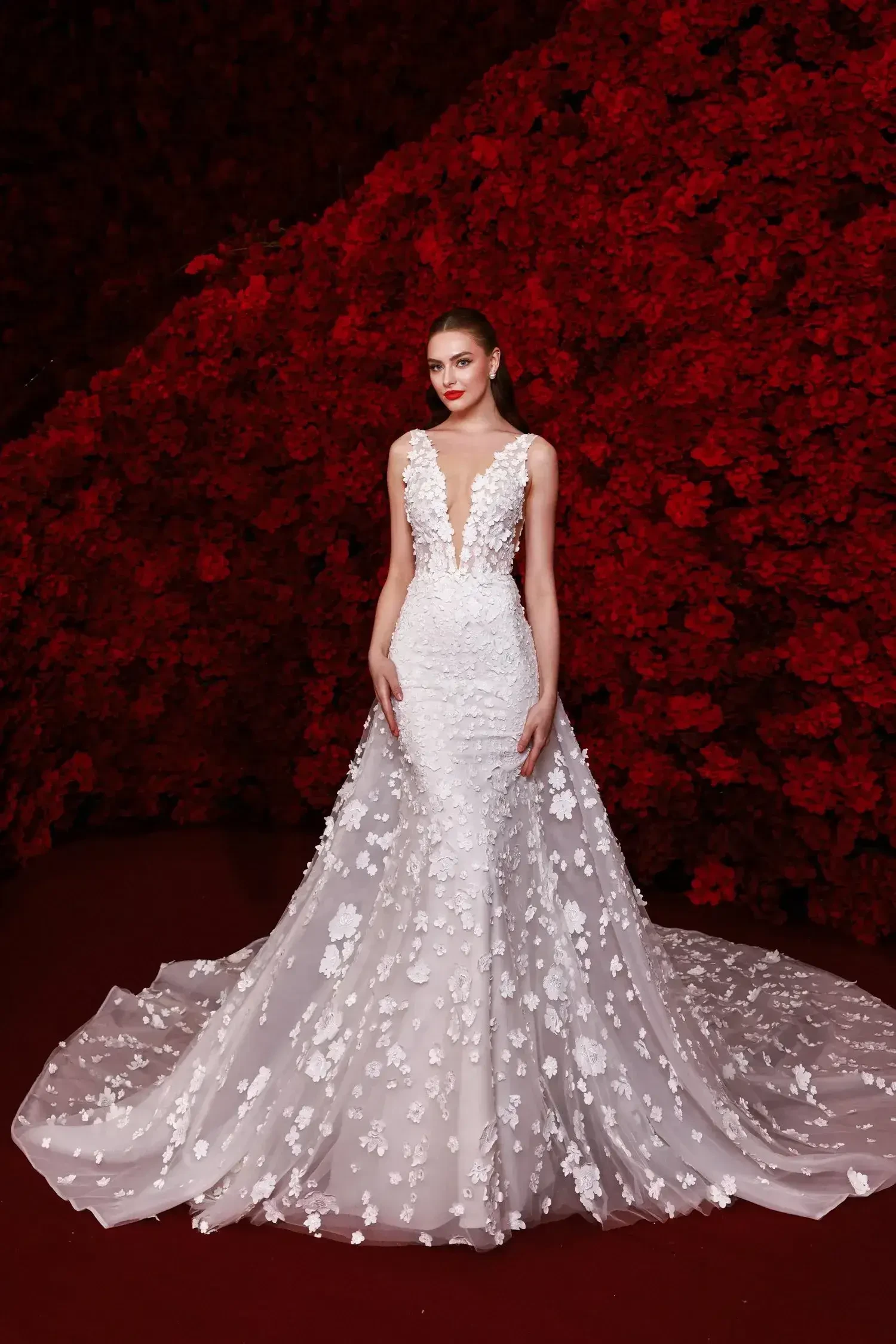 A woman in an elegant white gown adorned with floral designs, standing against a backdrop of red flowers.