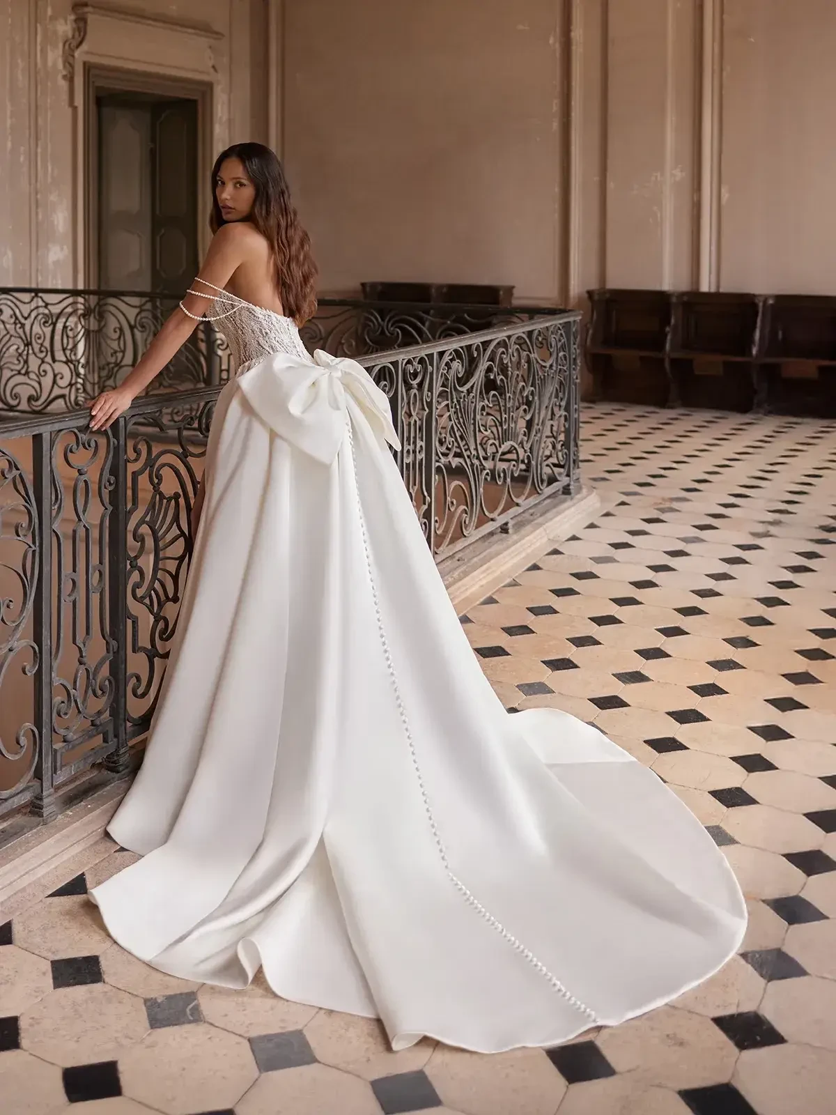 A woman in an elegant off-shoulder wedding gown with a long train and large bow stands on checkered floors beside ornate railings, evoking a timeless, romantic feel.