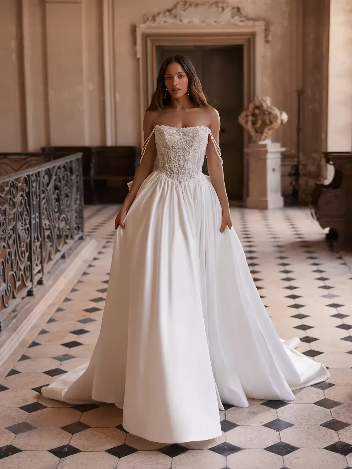 A woman in an elegant, strapless, white wedding gown with lace detail stands in a grand, historic hallway with intricate black railings and checkered floors.