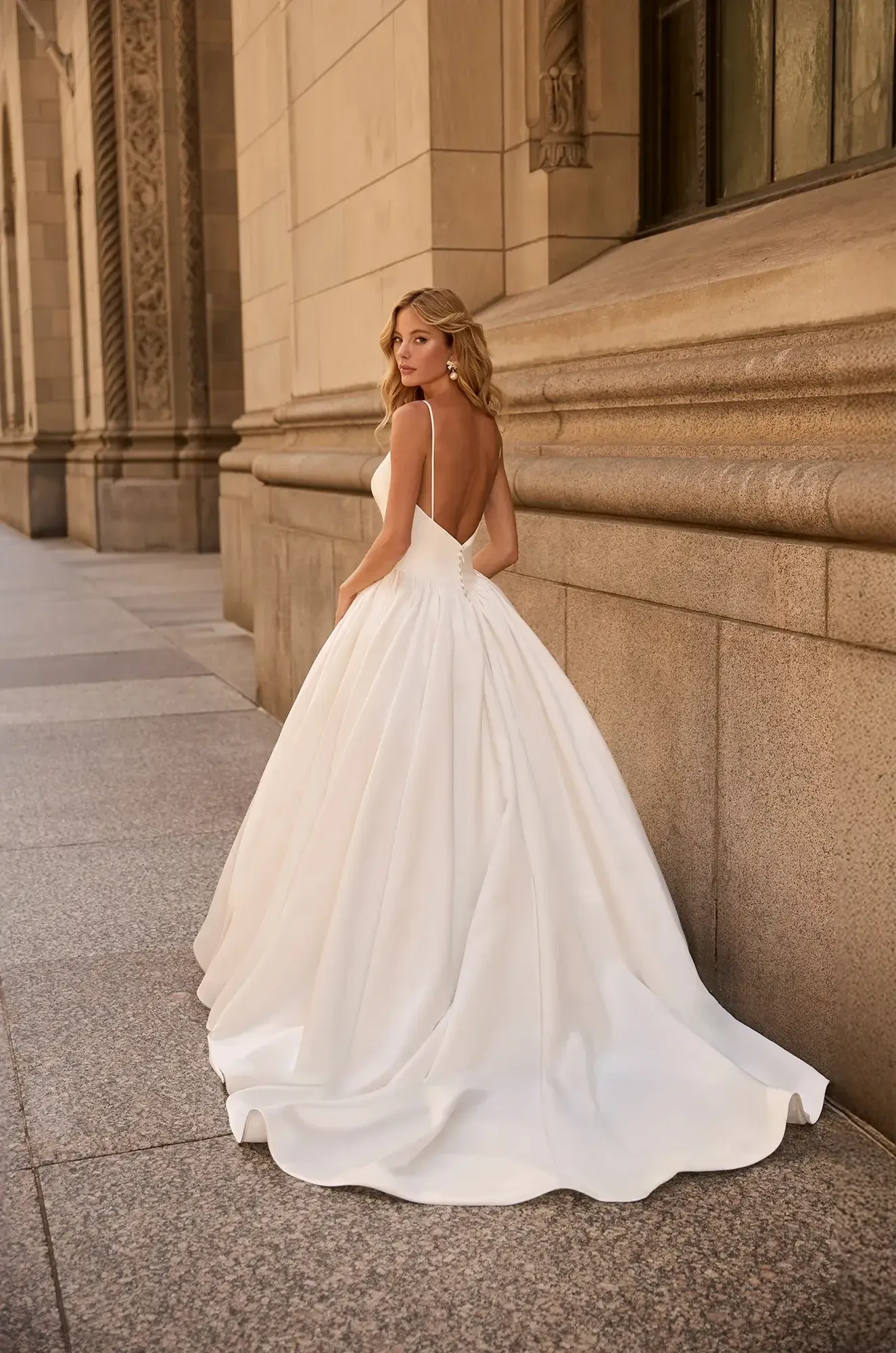 A woman in a backless, flowing white gown stands elegantly against a grand stone building, conveying a sense of timeless sophistication.