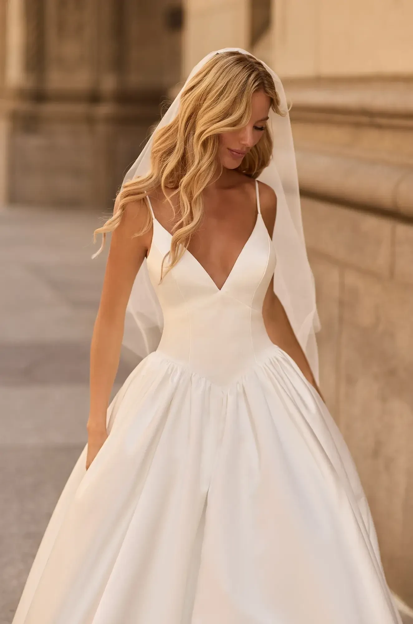 Bride in a white gown with a deep V-neck and full skirt, walking serenely outdoors. She has long, wavy hair, and wears a veil, exuding elegance.