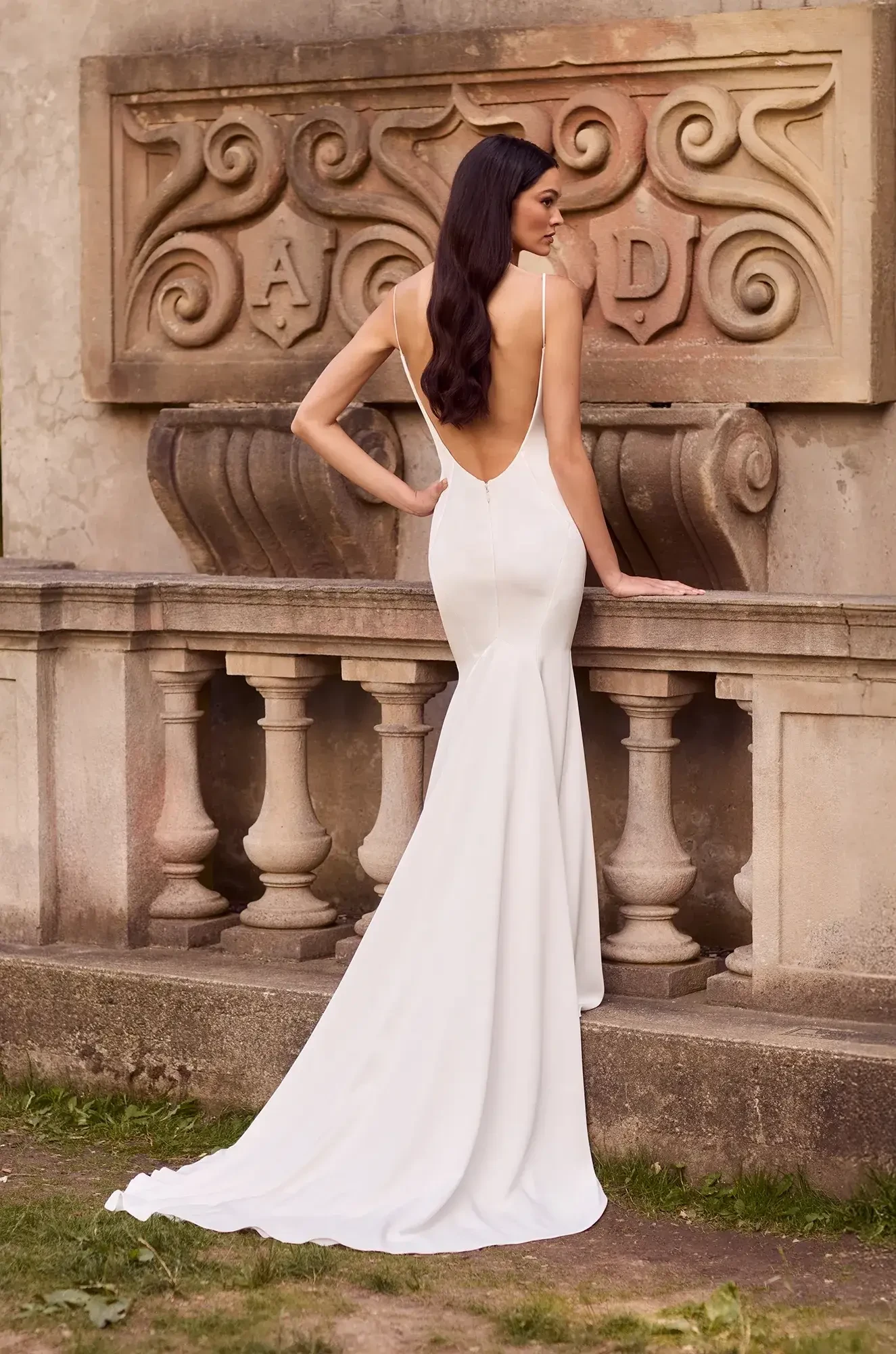 A model wearing a elegant white gown with a low back, posing by a decorative stone wall.